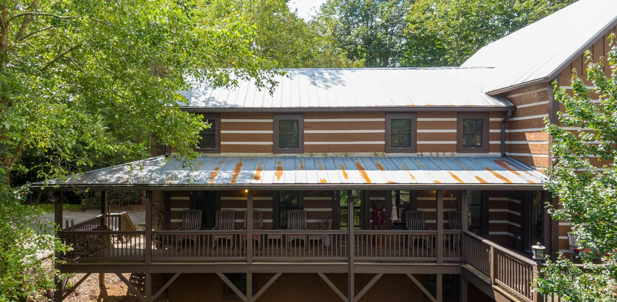 A rustic wooden porch with green foliage, metal roof, and striped wooden siding creates a warm and inviting entrance. The setting is perfect for unwinding in a rocking chair, sipping coffee, or gathering with family after a day outdoors.