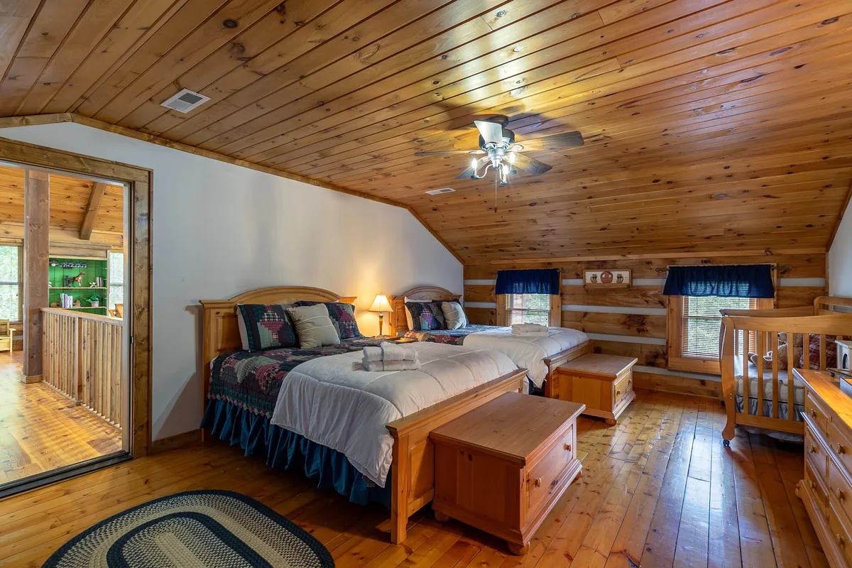 A family-friendly bedroom with two beds dressed in patterned bedspreads, wooden furniture, a window with blue curtains, and a ceiling fan. The room feels spacious and welcoming, providing ample space for guests or family members to share.