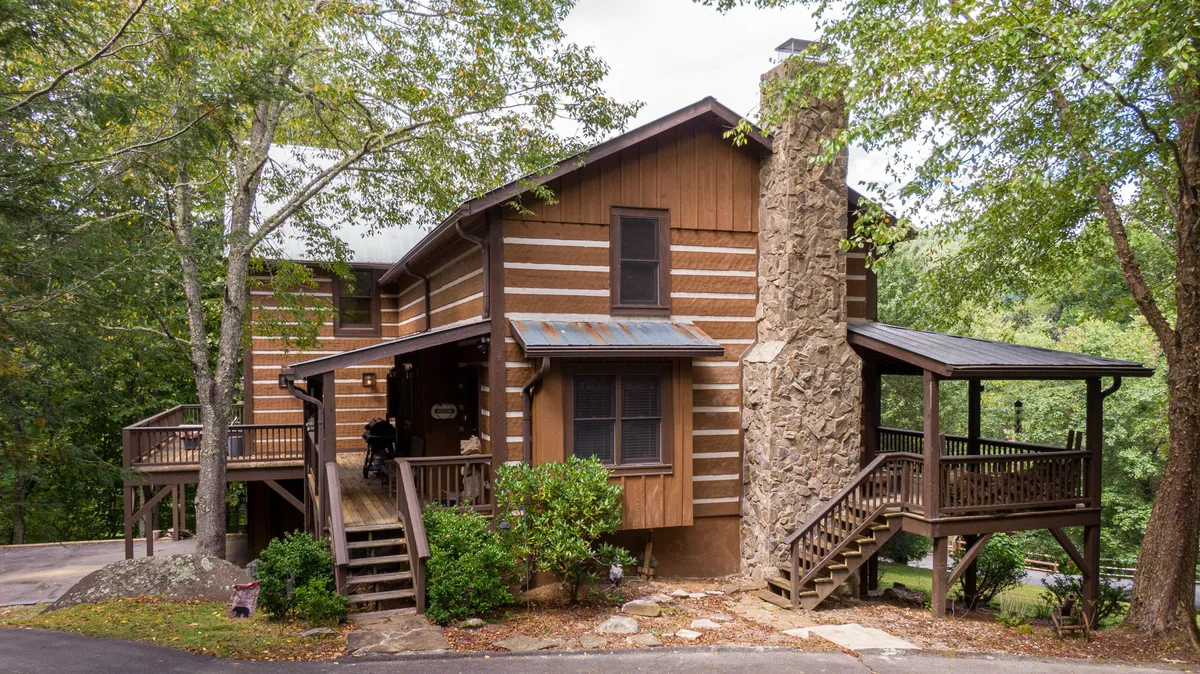 The image features a classic wooden log cabin-style home with a rustic stone chimney, surrounded by lush greenery. A spacious wooden deck offers views of the natural landscape, inviting guests to relax and take in the sunset. The scene evokes warmth and togetherness, perfect for family gatherings or cozy evenings by the fire.