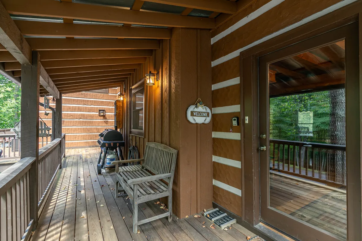 The image captures the welcoming exterior entrance, complete with a wooden bench, a friendly 'welcome' sign, a grill for outdoor cooking, and sturdy wooden beams. The scene suggests both hospitality and functionality, perfect for gatherings, barbecues, or simply enjoying the fresh air throughout the year.