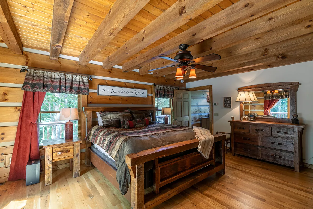 A serene bedroom features a large wooden bed, exposed rustic ceiling beams, a decorative mirror, and matching nightstands with lamps. The room is bathed in soft light, and the textures of wood and textiles create a calming, inviting atmosphere.