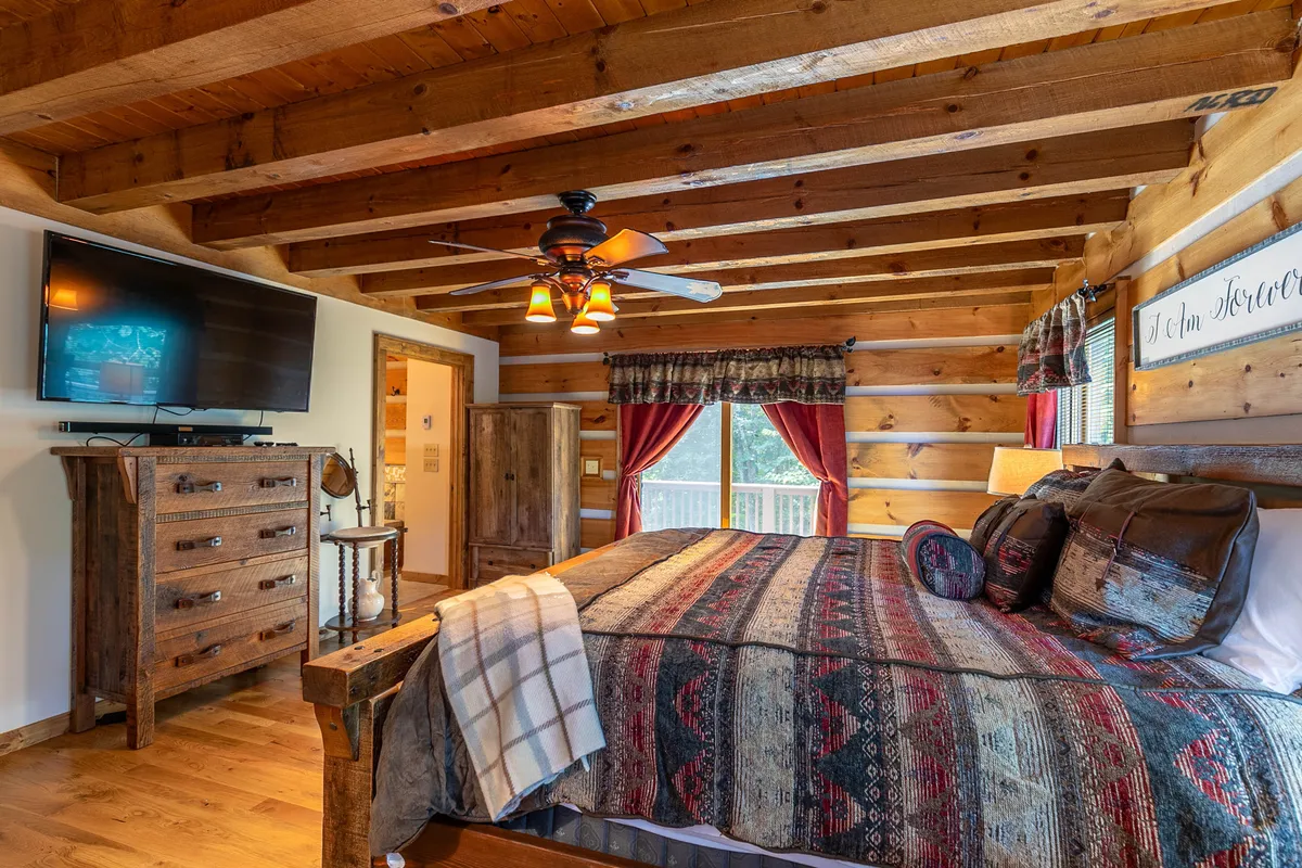 This image presents a bedroom with a wooden bed frame, plaid blanket, rustic dresser, and a large window that allows natural light to flood into the room. The décor is distinctly rustic, with warm wood tones and cozy textiles, and the presence of a TV adds a touch of modern comfort.