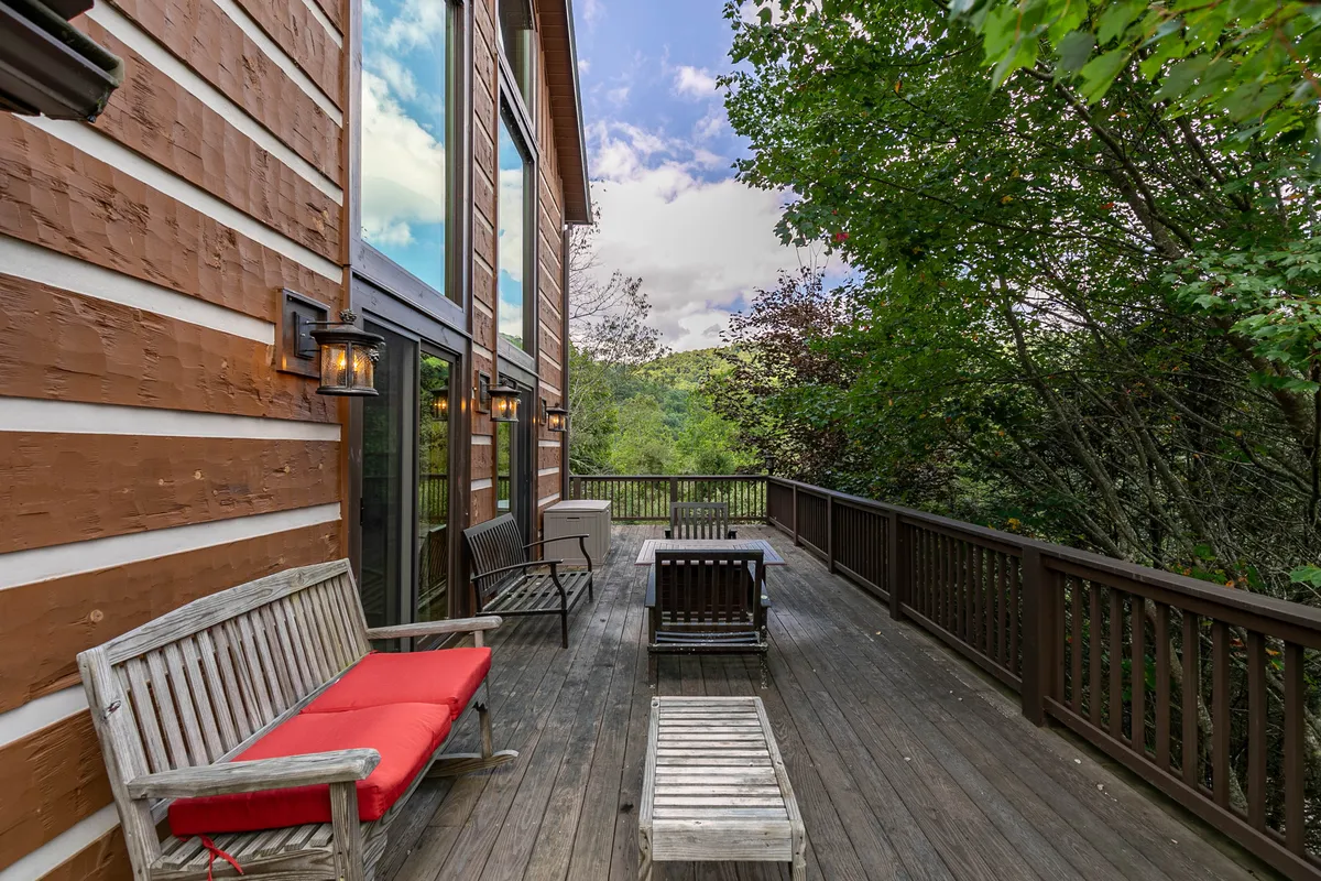 An inviting wooden deck with outdoor seating nestled amidst lush greenery. Large windows provide a seamless transition between the indoor living space and the tranquil outdoors. The setting is serene, perfect for relaxing, reading, or savoring a morning coffee while immersed in nature.
