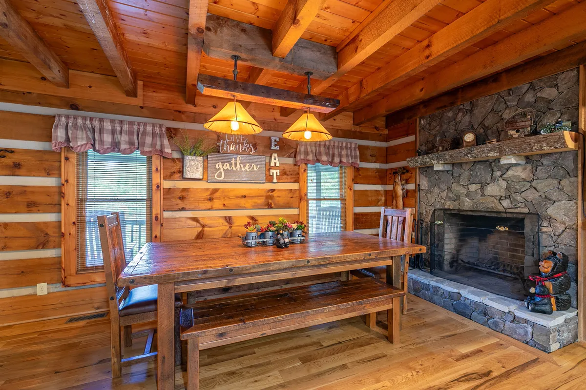 The adjacent dining area is anchored by a rustic wooden table, with a stone fireplace and exposed wooden beams creating a mountain lodge feel. Hanging lights and decorative wall signs add a warm, intimate touch, making the space perfect for shared meals and celebrations.