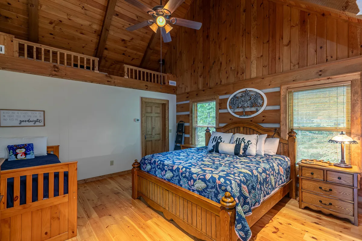 This inviting bedroom showcases a large wooden bed with decorative throw pillows, wooden furniture, a ceiling fan, and artwork on the wall. Natural light fills the room, creating a sense of openness and relaxation.