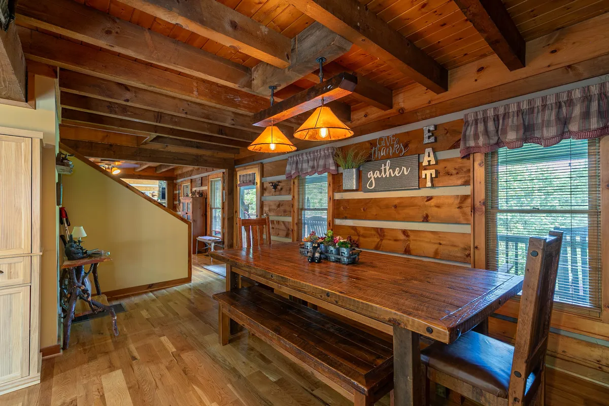 A large wooden dining table with hanging pendant lights overhead and decorative wall signs, set in a cozy, welcoming dining area. The table is expansive, ideal for group meals and lively conversation.