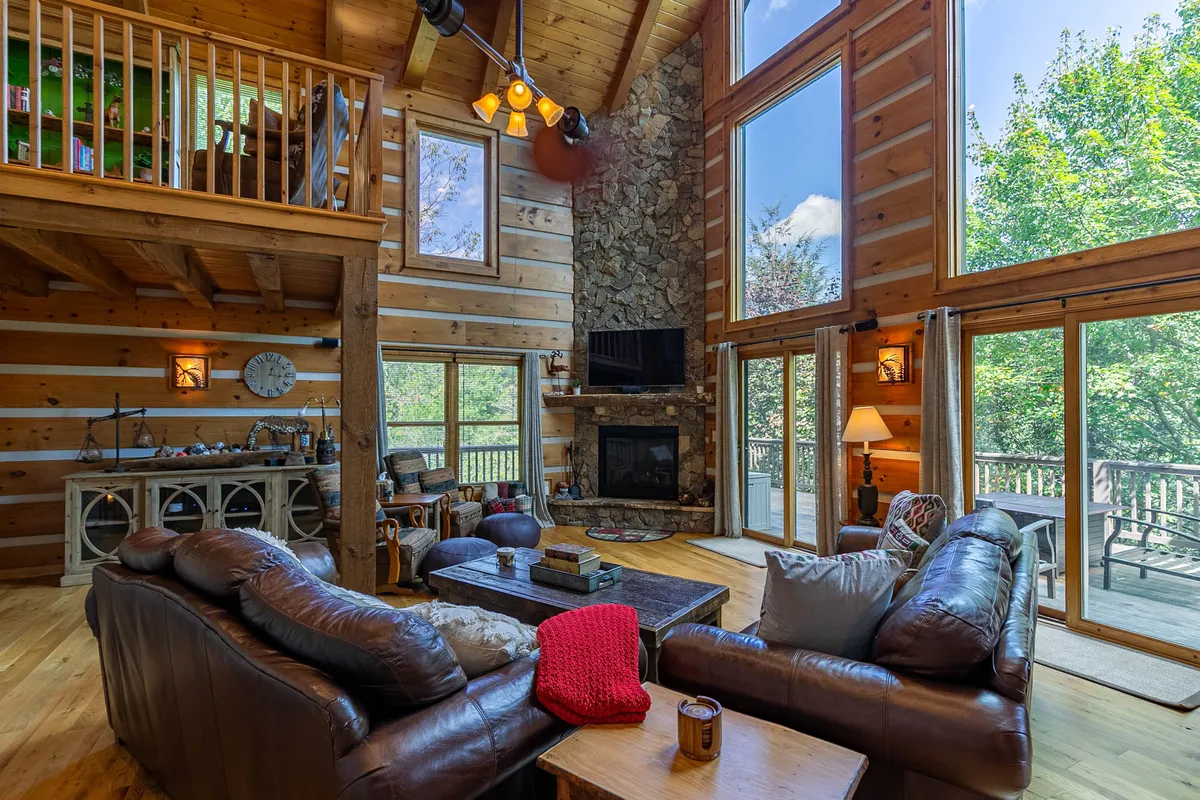 This image showcases a spacious open living area centered on a dramatic stone fireplace. Expansive windows flood the room with natural light, revealing woodland views. Comfortable seating is arranged to encourage conversation and relaxation, creating a welcoming atmosphere.