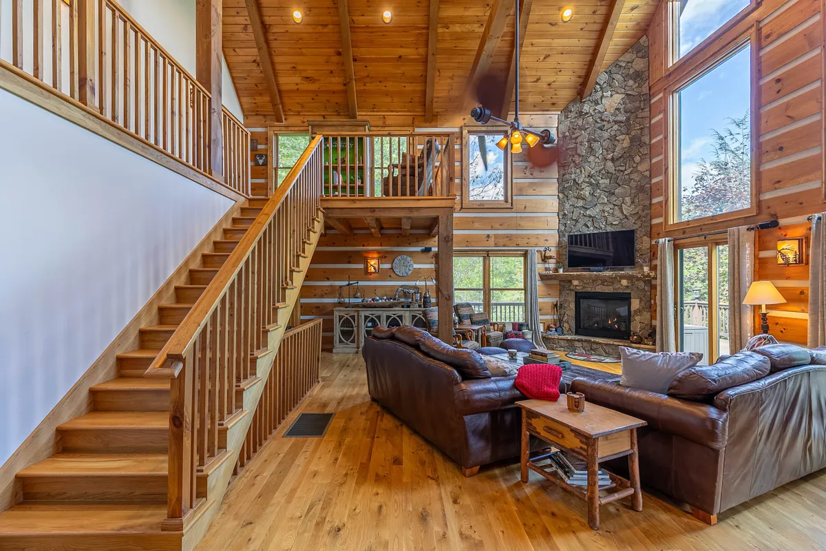 A soaring wooden ceiling frames an open-plan living space with a prominent stone fireplace, large windows with scenic forest views, and leather sofas arranged for gatherings. A wooden staircase adds rustic charm, while warm lighting enhances the atmosphere.