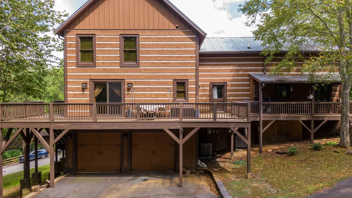 A detailed exterior shot of the log cabin, emphasizing the wooden deck, large windows, and garage doors. The deck offers ample space for outdoor dining or simply enjoying the natural setting, reinforcing the connection between the home and its surrounding wilderness.